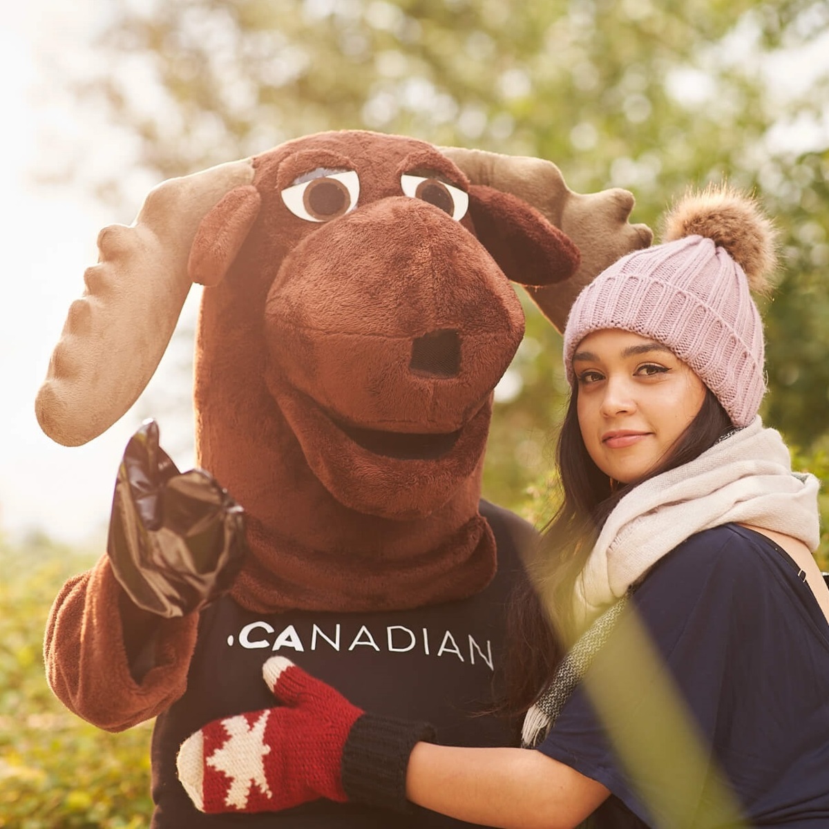 A photo of a person in a moose costume being hugged by a lady in a touque and gloves. It's _not_ winter